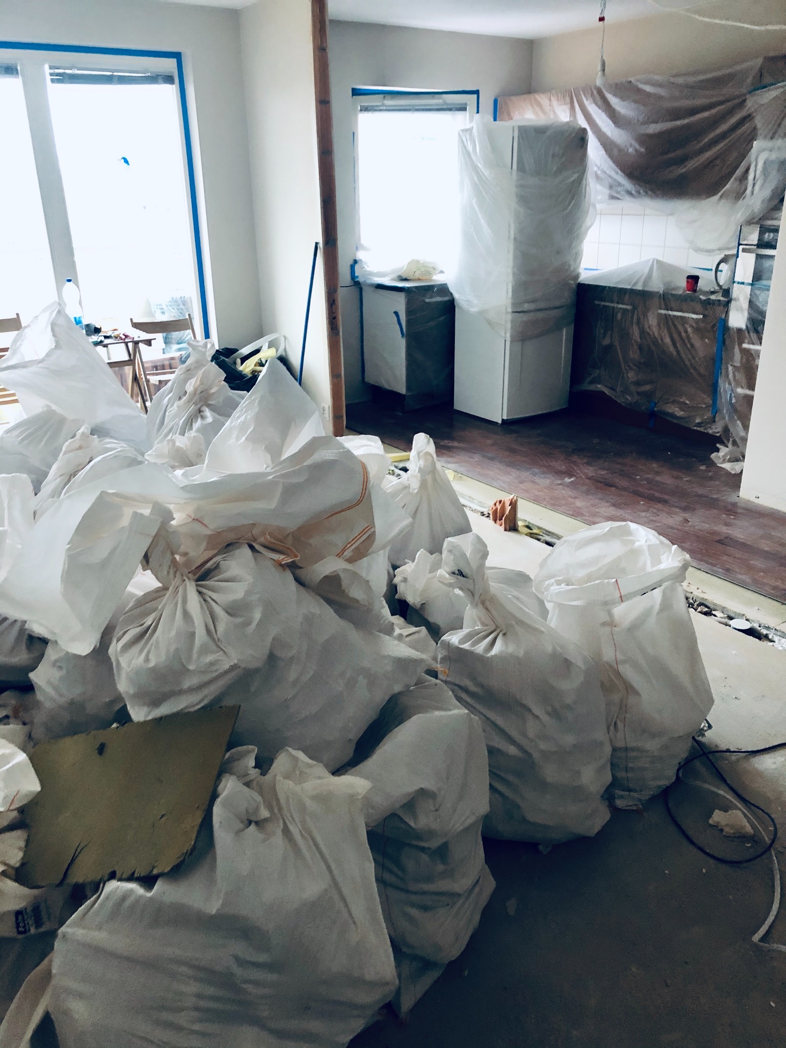 Stos worków z odpadami po remoncie kuchni, widoczne zabezpieczone folią meble kuchenne i okna z niebieską taśmą malarską.