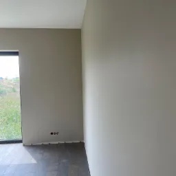 Wnętrze pokoju po świeżym pomalowaniu ścian na beżowo-szary odcień, z widocznymi trzema otworami na gniazdka elektryczne i ułożoną ciemną podłogą panelową, światło dzienne wpadające przez okno.