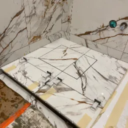 Montaż płytek imitujących marmur w kabinie prysznicowej z odpływem liniowym, widoczne klipsy poziomujące i taśma zabezpieczająca.