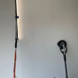 Świeżo wyszpachlowana ściana oświetlona lampą inspekcyjną, obok szlifierka do gładzi z podłączonym odkurzaczem, widoczny niedokończony dolny fragment ściany.