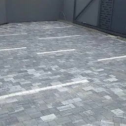 Ułożona szara kostka brukowa z białymi liniami, stos kostki obok czerwonego słupka, szara brama w tle.