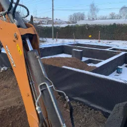 Pomarańczowe ramię koparki na tle częściowo wykopanych fundamentów z bloczków betonowych, zabezpieczonych folią kubełkową, zasypanych ziemią, w zimowej scenerii.