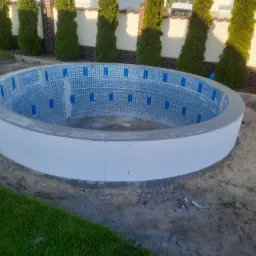 Okrągły basen w trakcie budowy, wyłożony niebieską mozaiką z zabezpieczającą folią, otoczony trawnikiem i tujami.