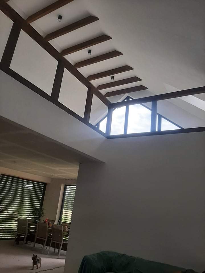 Wysoki sufit z drewnianymi belkami i oknem dachowym w jasnym wnętrzu, widoczny fragment jadalni z żaluzjami i mały pies.