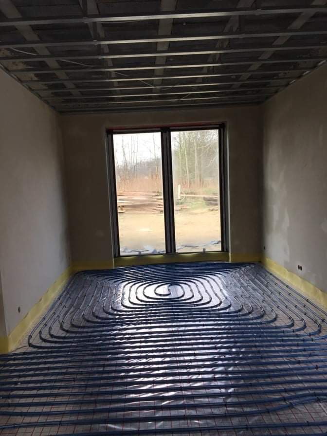 Niedokończone pomieszczenie z ułożonymi rurami ogrzewania podłogowego na folii, widoczne taśmy zabezpieczające ściany i sufit podwieszany w trakcie montażu, okno z widokiem na zewnątrz.