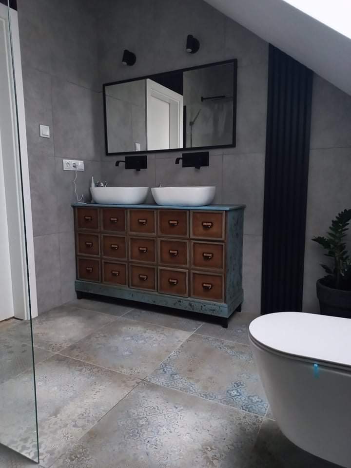 Szara łazienka na poddaszu z umywalkami nablatowymi na komodzie z szufladami, czarnymi lampami i grzejnikiem żeberkowym w pionie.