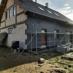 Dom jednorodzinny w trakcie montażu izolacji termicznej elewacji, widoczne rusztowania, częściowo ocieplona ściana zewnętrzna styropianem grafitowym oraz fragment ściany z bloczków silikatowych,...