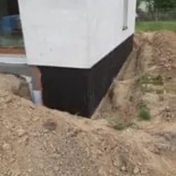 Fundament budynku z widoczną izolacją przeciwwilgociową w kolorze czarnym, częściowo zasypany ziemią, z wykopem odsłaniającym rurę odpływową. W tle trawa i fragment budynku sąsiedniego.