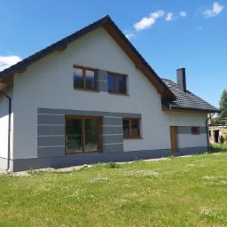 Dwukondygnacyjny dom jednorodzinny z jasną elewacją, brązowymi oknami i szarymi akcentami dekoracyjnymi, widoczny z perspektywy trawnika w słoneczny dzień.