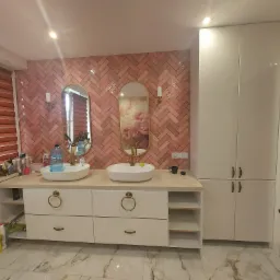 Łazienka z dwiema owalnymi umywalkami nablatowymi, r&oacute;żową ceglaną ścianą ułożoną w jodełkę, złotymi lustrami i białymi szafkami z okrągłymi uchwytami, widoczna butelka wody mineralnej.