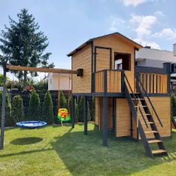 Drewniany domek dla dzieci na podwyższeniu z drabinką i huśtawką na tle zadbanego trawnika i nowoczesnego domu.