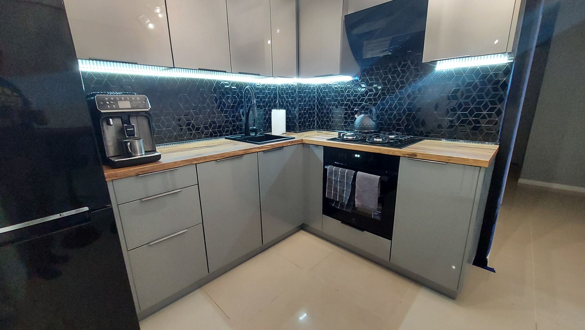 Nowoczesna kuchnia w szarościach z drewnianym blatem, geometryczną mozaiką na ścianie i oświetleniem LED pod szafkami, widoczny ekspres do kawy i piekarnik z ręcznikami.