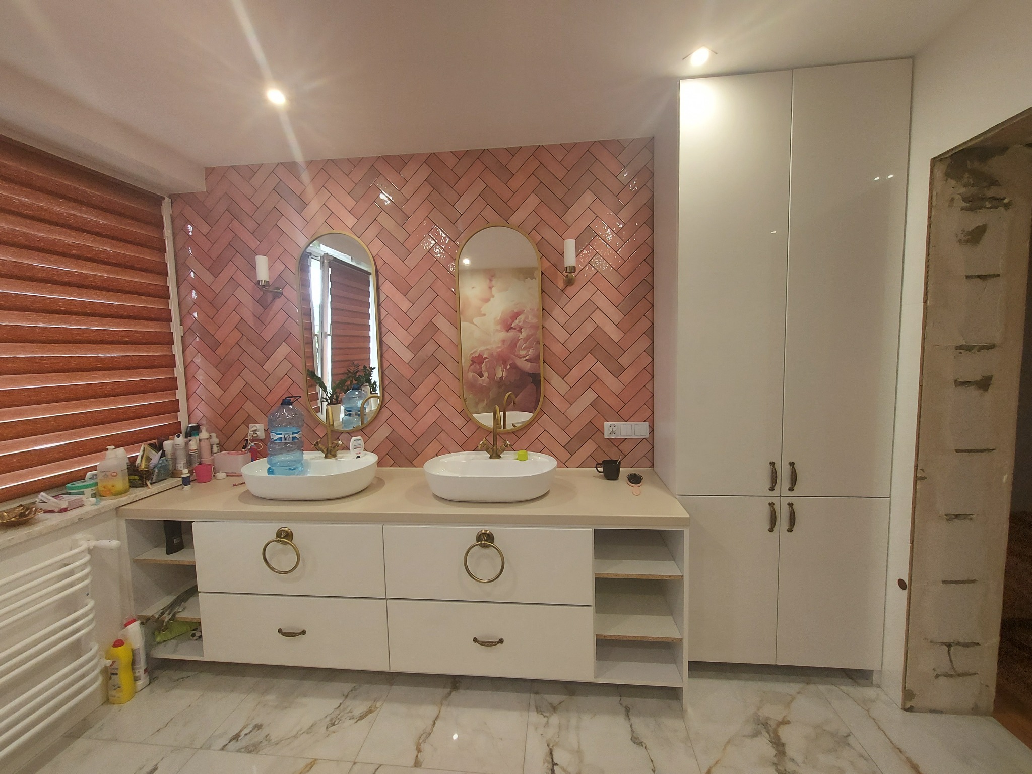 Łazienka z dwiema owalnymi umywalkami nablatowymi, różową ceglaną ścianą ułożoną w jodełkę, złotymi lustrami i białymi szafkami z okrągłymi uchwytami, widoczna butelka wody mineralnej.