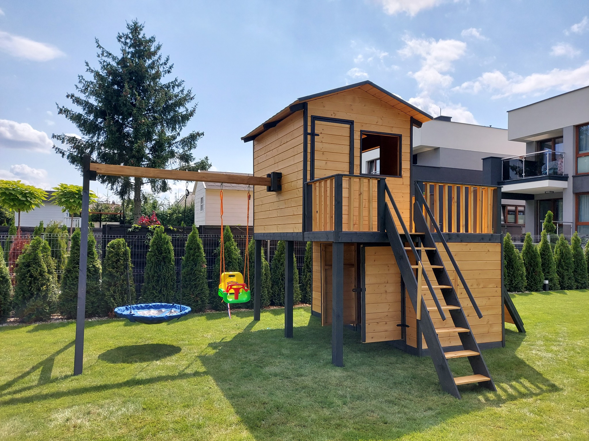 Drewniany domek dla dzieci na podwyższeniu z drabinką i huśtawką na tle zadbanego trawnika i nowoczesnego domu.