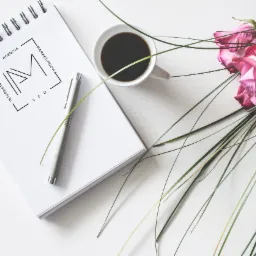 Notatnik z logo Agencji Marketingowej, długopis, filiżanka kawy i różowe róże na białym tle.