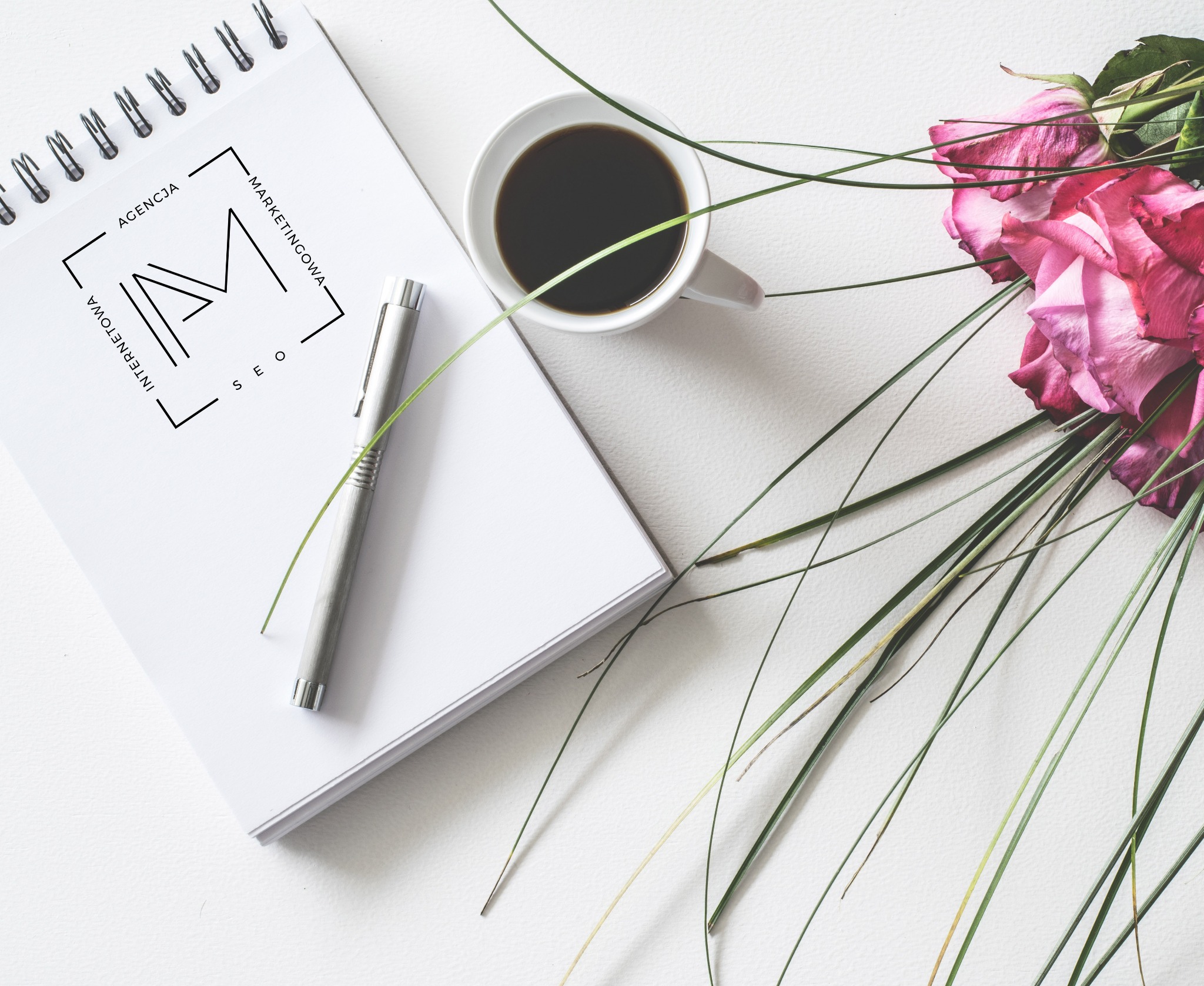 Notatnik z logo Agencji Marketingowej, długopis, filiżanka kawy i różowe róże na białym tle.