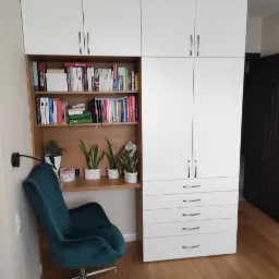 Biała szafa wnękowa z biurkiem i regałem na książki, uzupełniona zielonym krzesłem i roślinami doniczkowymi, na podłodze panele.