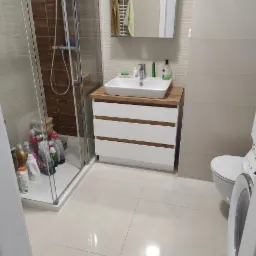 Nowoczesna łazienka z białą szafką pod umywalką z drewnianym blatem, kabiną prysznicową z brązowymi płytkami i toaletą. Widoczne kosmetyki i środki czystości.