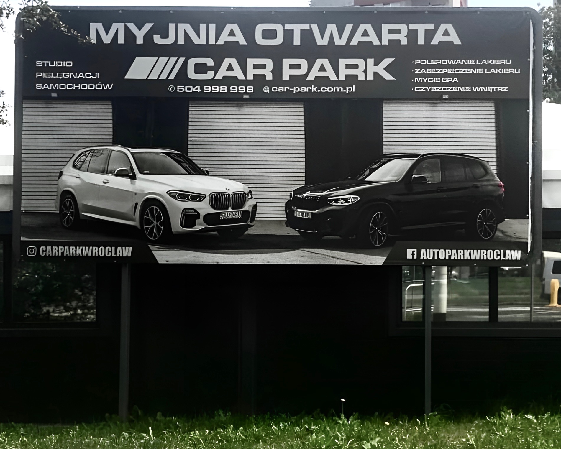 Banner reklamowy myjni samochodowej z dwoma samochodami BMW, biały i czarny, na tle zamkniętych bram garażowych.