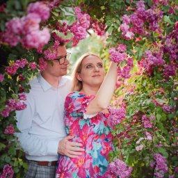 Gocha Chmiel Fotografia i Film - Zakochana para w kwitnącym ogrodzie róż. Kobieta w kwiecistej sukience sięga po kwiat. Mężczyzna w okularach obejmuje ją czule. Romantyczna sesja w plenerze.