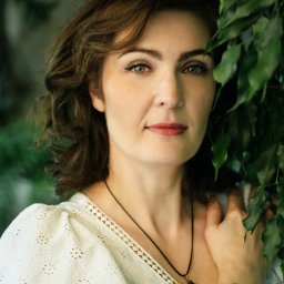 Gocha Chmiel Fotografia i Film - Portret kobiety o zielonych oczach, z delikatnym uśmiechem, w białej bluzce z koronką, pozującej na tle zielonych liści. Subtelna biżuteria dodaje elegancji.