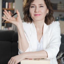 Gocha Chmiel Fotografia i Film - Portret kobiety w białym żakiecie, trzymającej długopis, opierającej się o stos książek na tle regału. Profesjonalne oświetlenie, ciepłe barwy, elegancki wygląd.