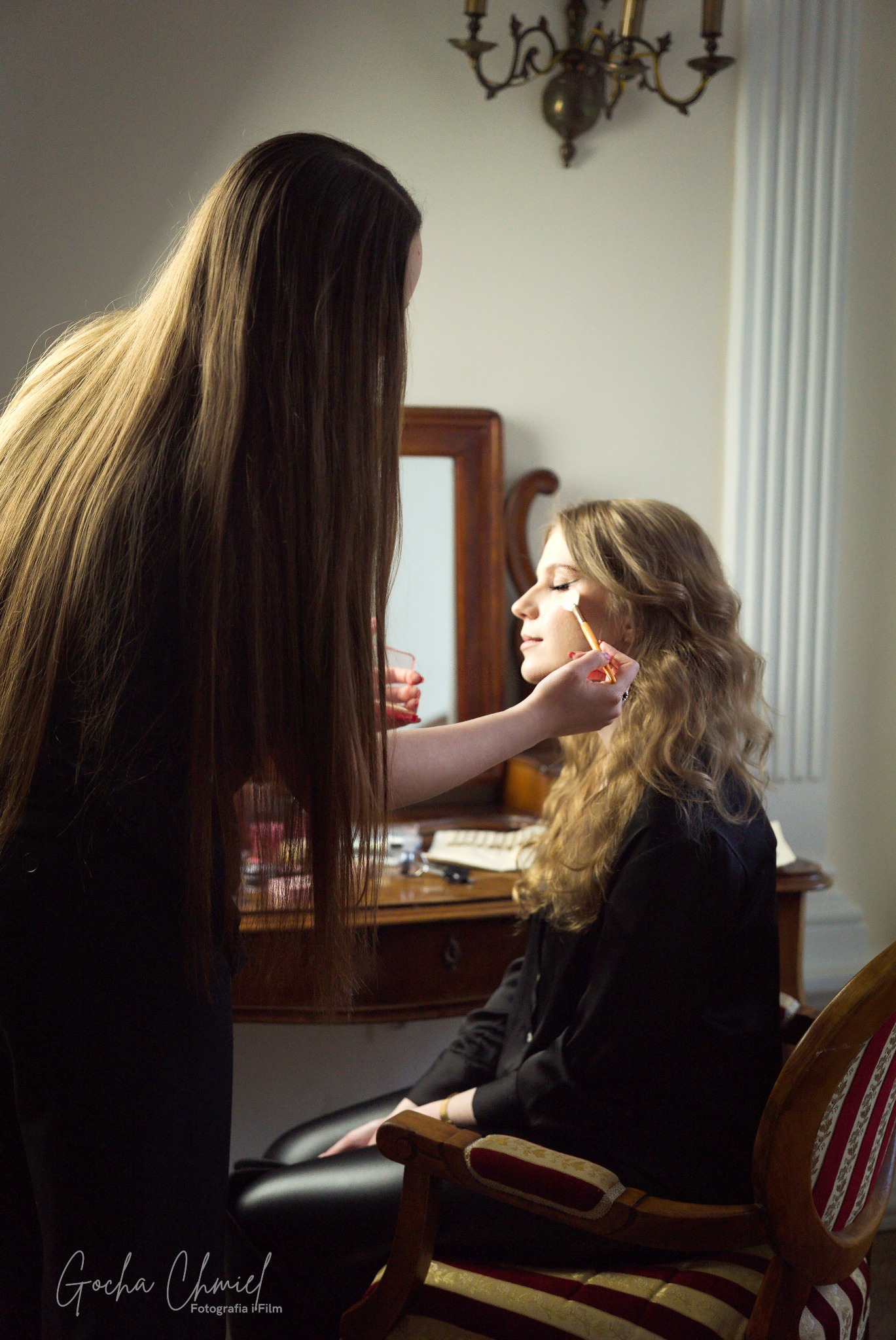 Wizażystka z długimi, brązowymi włosami nakłada pędzlem makijaż na powiekę jasnowłosej kobiety siedzącej na krześle w eleganckim wnętrzu w Kielcach.