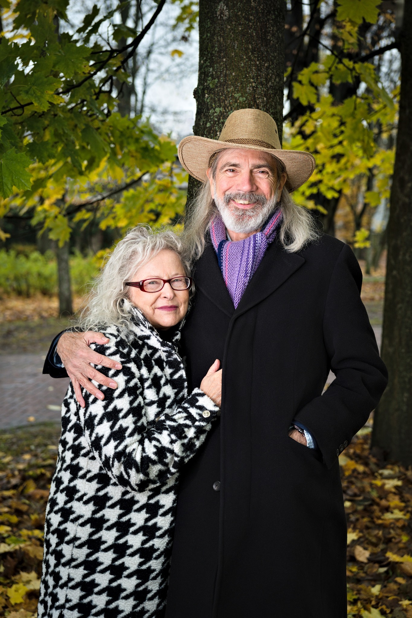 Uśmiechnięta para seniorów obejmująca się na tle jesiennego parku: kobieta w czarno-białym płaszczu w pepitkę i mężczyzna w kapeluszu i czarnym płaszczu, liście na ziemi.