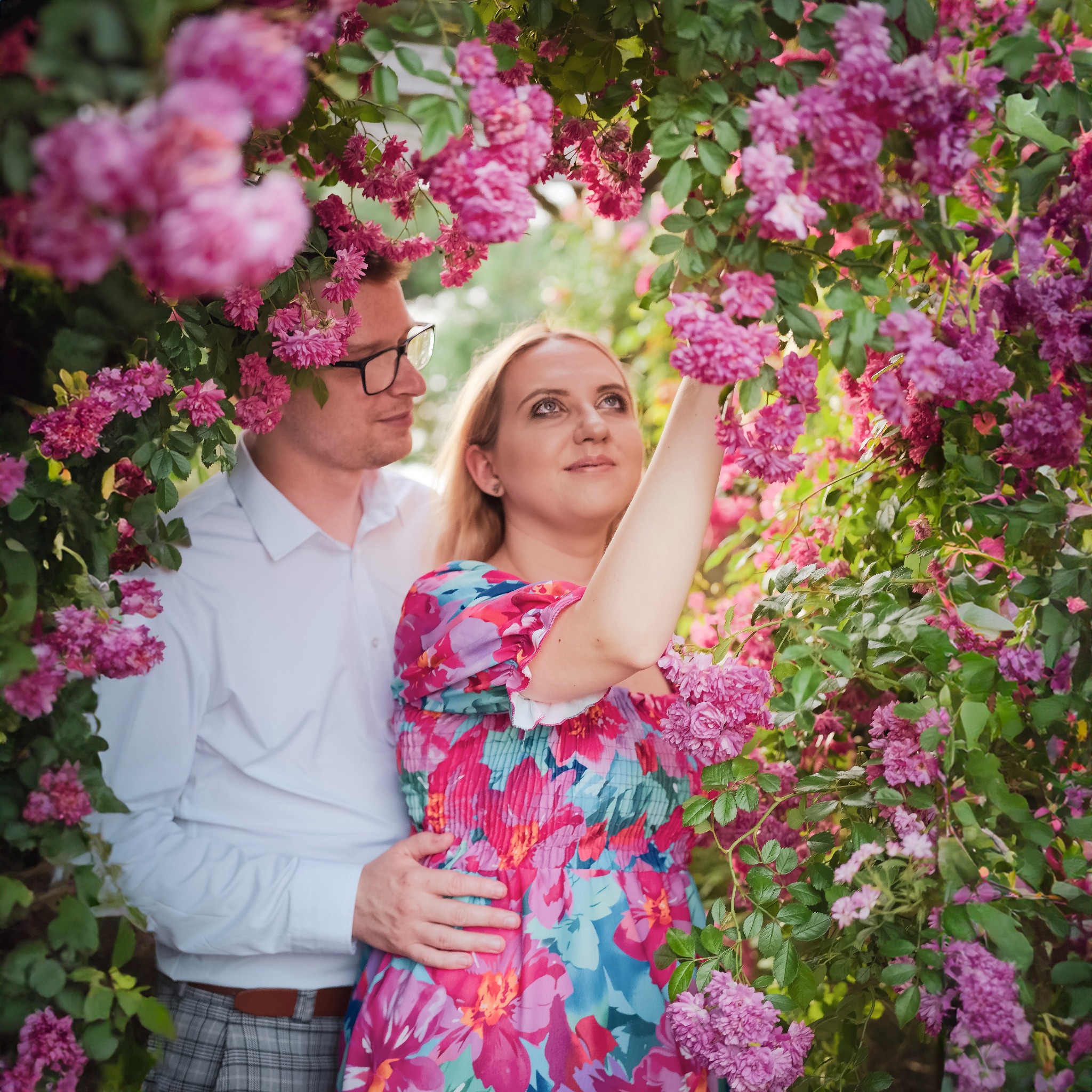 Zakochana para w kwitnącym ogrodzie róż. Kobieta w kwiecistej sukience sięga po kwiat. Mężczyzna w okularach obejmuje ją czule. Romantyczna sesja w plenerze.