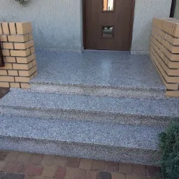 Zewnętrzne schody wejściowe z szarego, nakrapianego kamienia prowadzące do drzwi, z ceglanymi filarami po bokach i brązową skrzynką na listy.