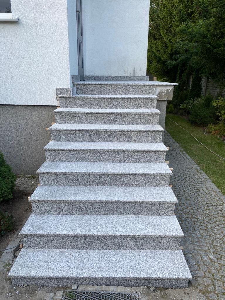 Zewnętrzne schody z szarego granitu prowadzące do drzwi wejściowych domu, widoczne detale wykończenia i kostka brukowa obok.
