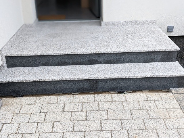 Dwu-stopniowe schody wejściowe z jasnoszarego kamienia z ciemnoszarymi podstopnicami, prowadzące do drzwi wejściowych, widok z góry w lekkim oświetleniu.