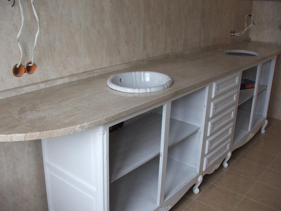 Beżowy blat z kamienia z wpuszczoną białą umywalką, biała szafka pod umywalkę w stylu retro z szufladami i półkami, widoczne wystające kable zasilające w ścianie.