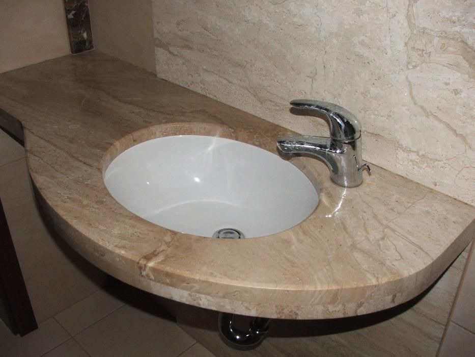 Umywalka wpuszczona w beżowy blat z kamienia naturalnego o zaokrąglonym kształcie, chromowana bateria i odpływ, dopasowana kolorystycznie okładzina ściany.