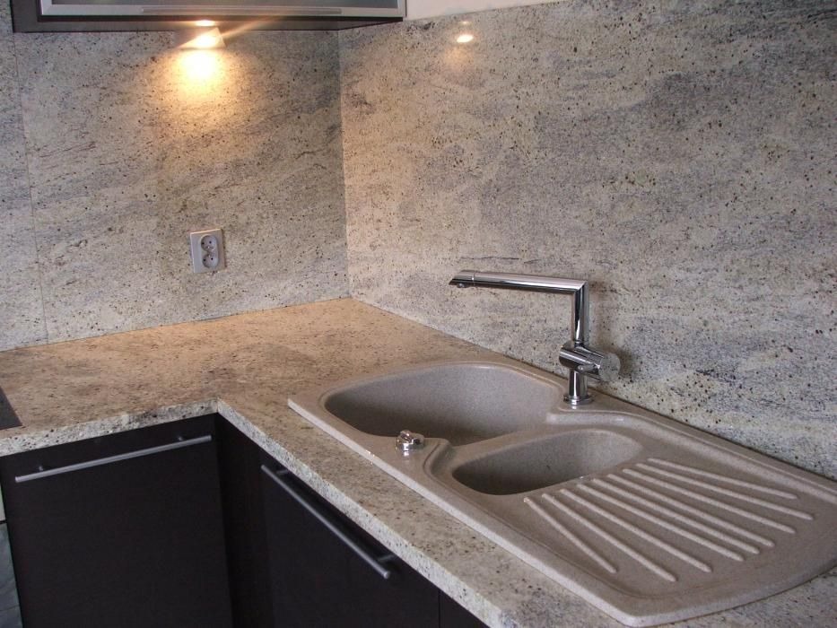 Kuchnia z blatem z jasnego kamienia o delikatnym wzorze, dopasowanym do ściany nad zlewem; chromowana bateria i ciemne szafki z podłużnymi uchwytami.