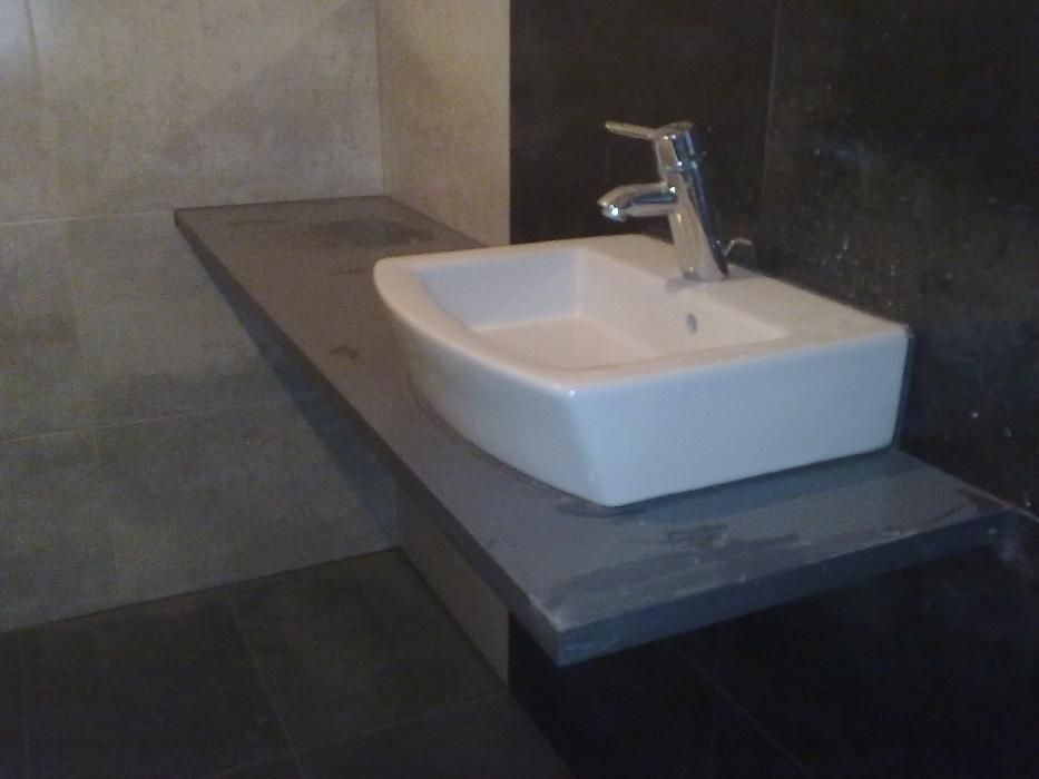 Biała, prostokątna umywalka nablatowa z chromowaną baterią, osadzona na szarym blacie w łazience z ciemną ścianą.