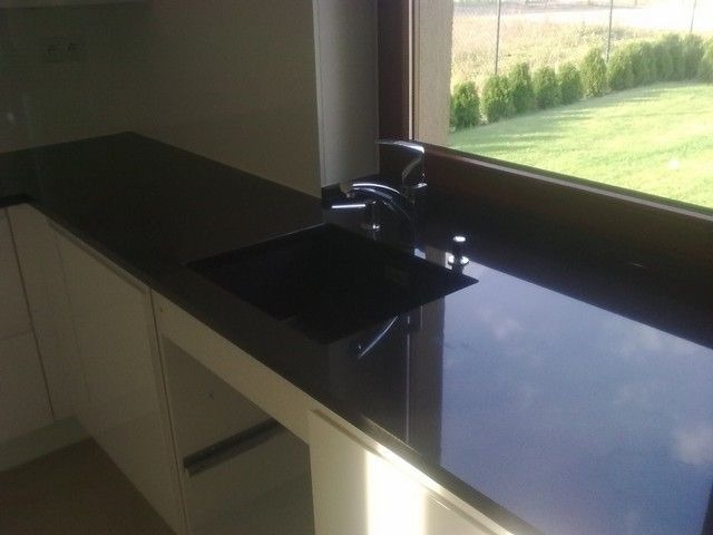 Błyszczący, czarny blat kamienny przy oknie z widokiem na trawnik, nowoczesna kuchnia z białymi szafkami.