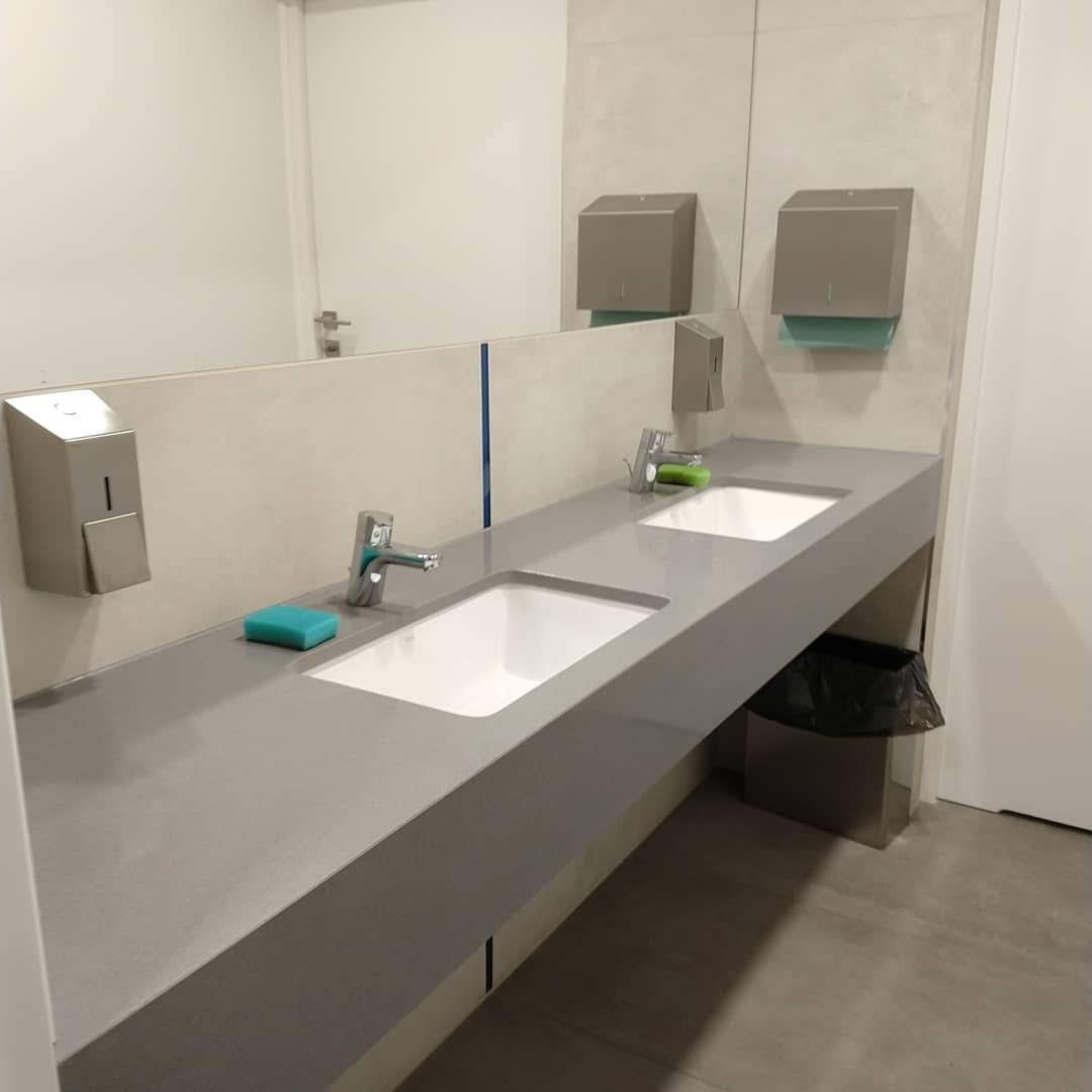 Długa, szara umywalka z dwoma białymi zlewami i bateriami, z dozownikiem mydła i ręczników papierowych w publicznej toalecie.