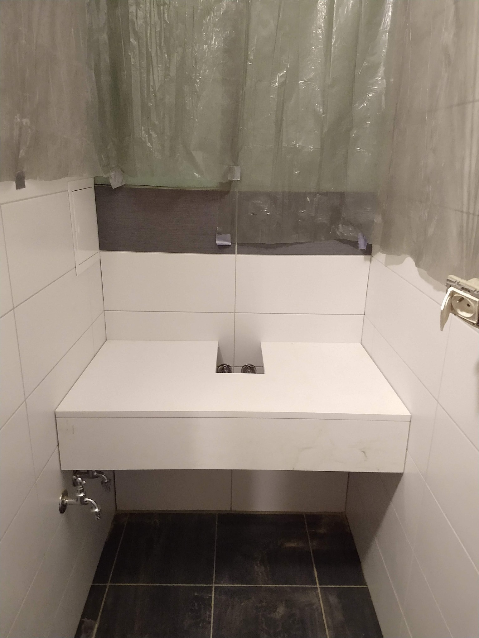 Biały blat umywalkowy z dwoma otworami na baterie, zamontowany w łazience z białymi płytkami i ciemną podłogą, zabezpieczony folią malarską.