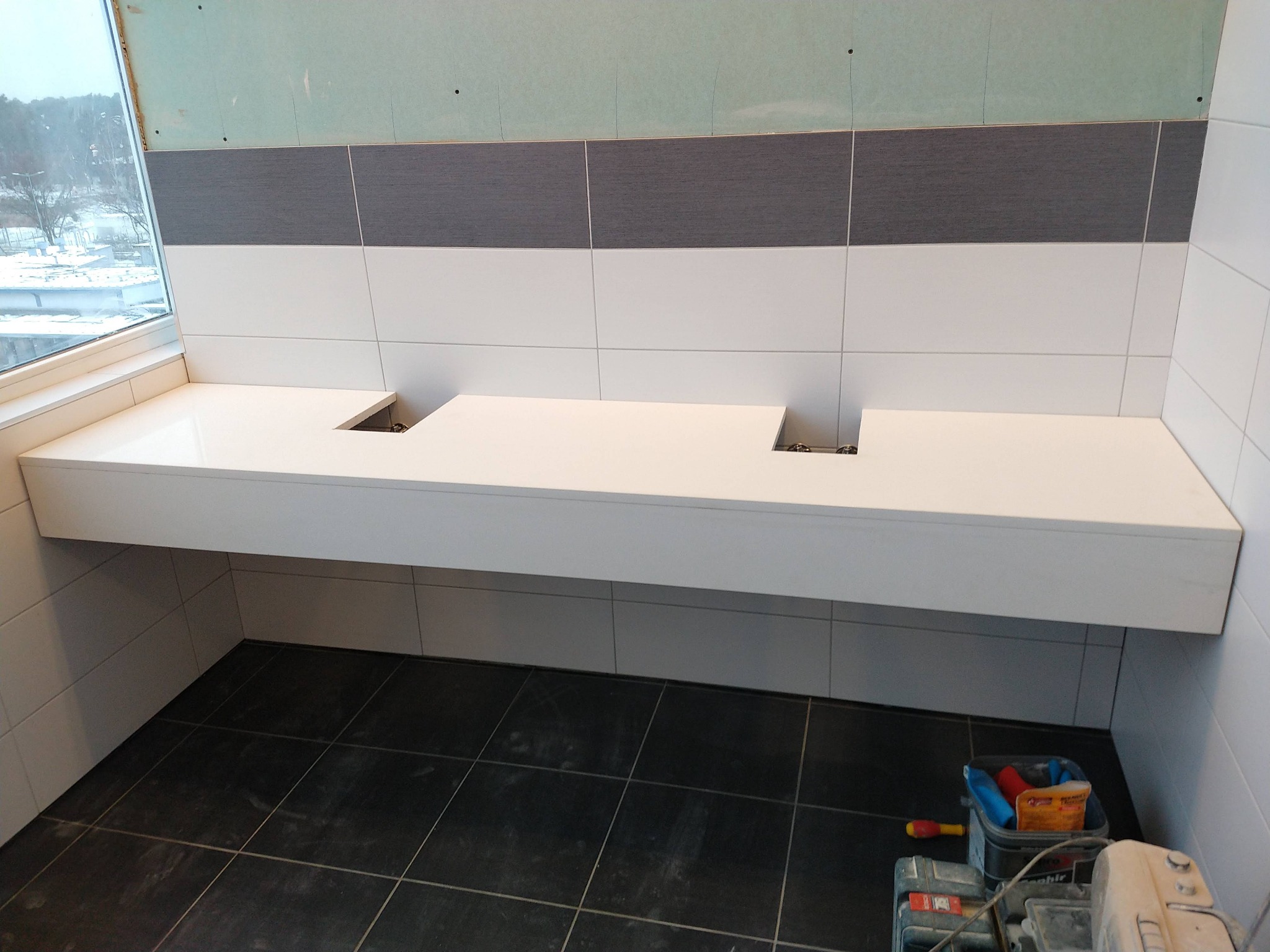 Biały, wiszący blat z dwoma otworami na umywalki, zamontowany w łazience z szaro-białymi płytkami, widoczne narzędzia pracy.