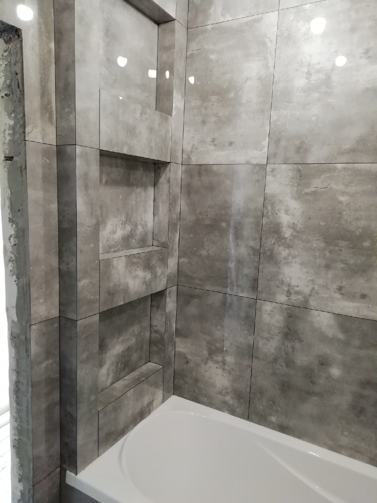 Wykończona łazienka z szarymi, marmurkowymi płytkami i wnękami w ścianie nad białą wanną.