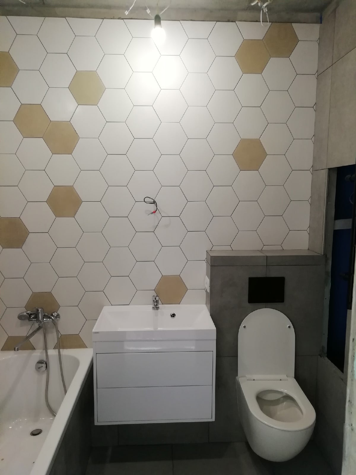Wykończona łazienka z heksagonalnymi białymi i złotymi płytkami na ścianie, białą szafką z umywalką, wanną z baterią i toaletą z szarą zabudową.