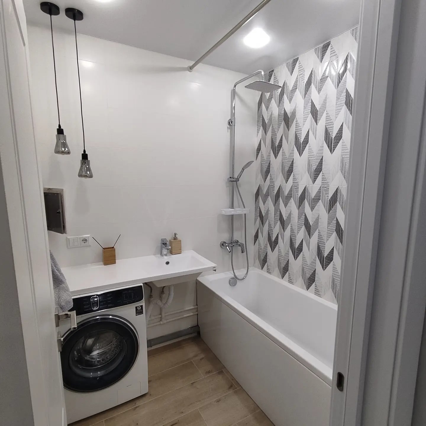 Nowoczesna łazienka z białą armaturą, wzorzystymi płytkami pod prysznicem i pralką pod umywalką.