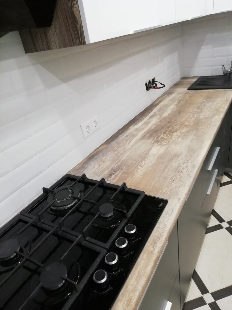 Czarna płyta gazowa z czterema palnikami w nowoczesnej kuchni z białymi płytkami i blatem o teksturze drewna, widoczne gniazdka elektryczne i fragment instalacji.