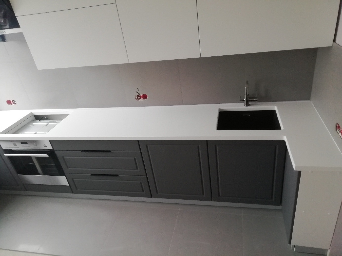 Nowoczesna kuchnia z szarymi szafkami, białym blatem i czarnym zlewozmywakiem; widoczne wystające kable elektryczne przygotowane do podłączenia urządzeń.