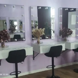 Trzy stanowiska do makijażu z lustrami podświetlanymi żarówkami, białymi blatami i czarnymi krzesłami barowymi na tle fioletowej ściany.