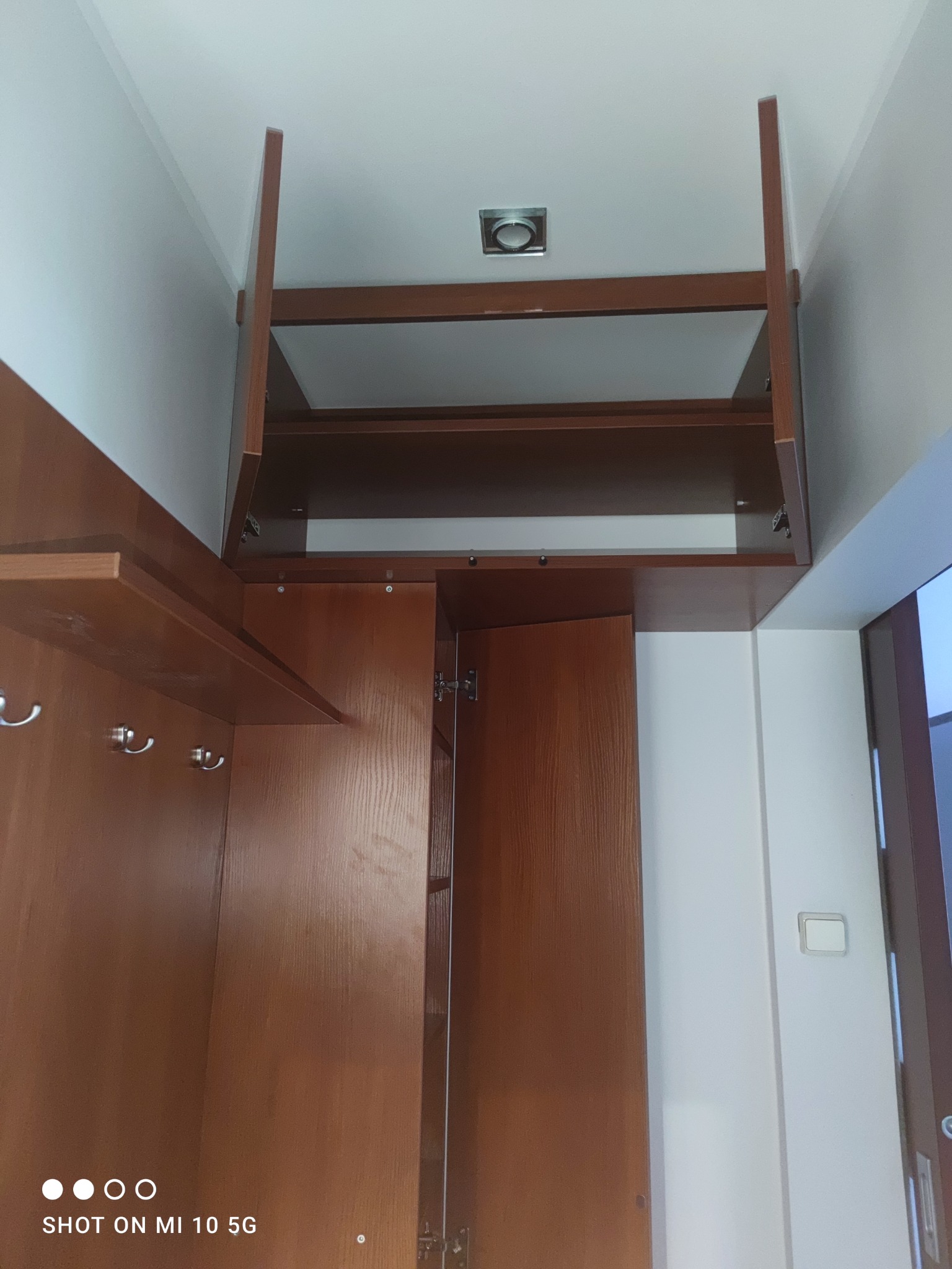 Wysoka szafa wnękowa z drewna w kolorze orzecha, z otwartą górną szafką i widocznymi haczykami na ubrania, zamontowana pod sufitem z kwadratowym oświetleniem.
