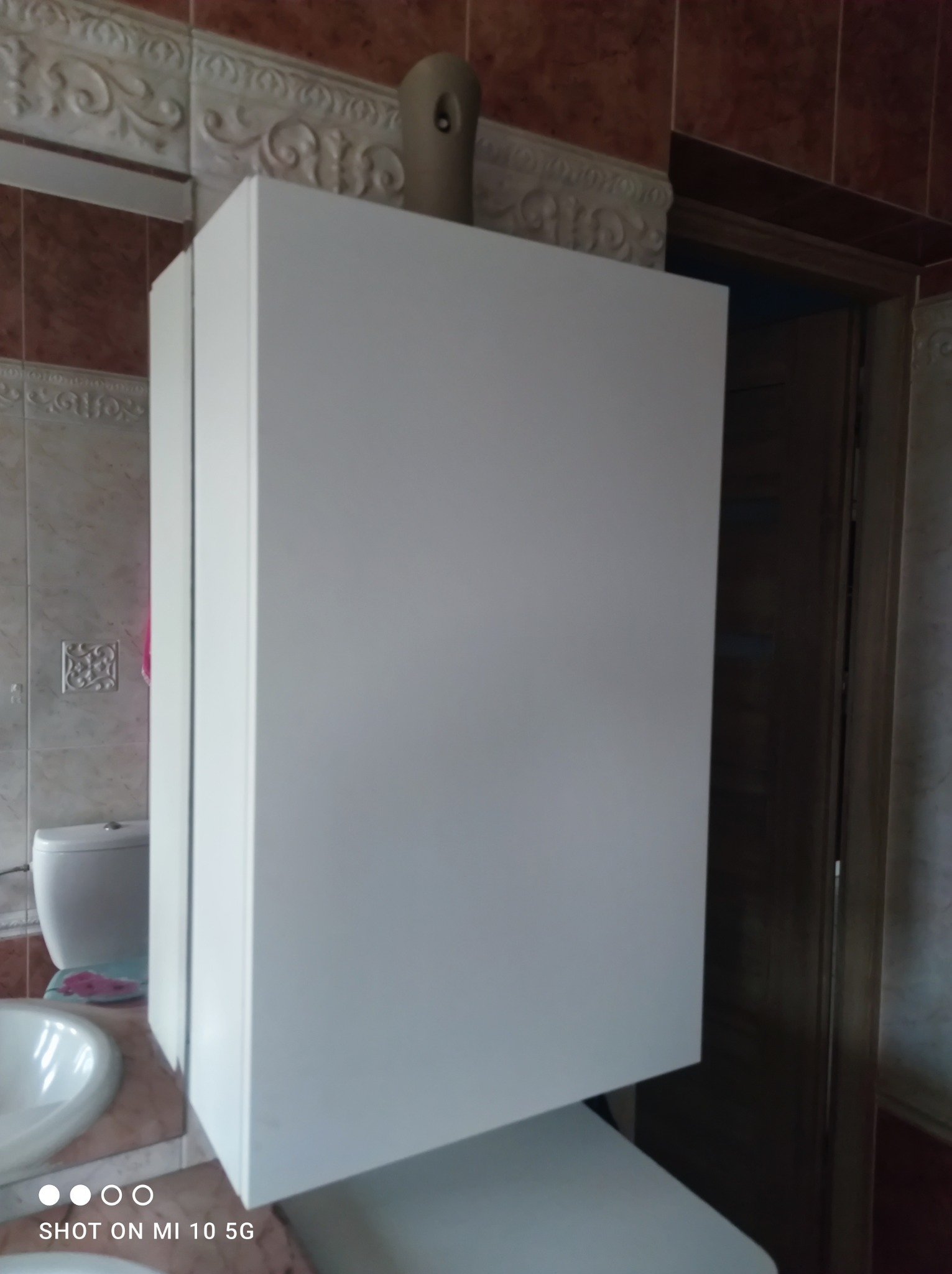 Biała szafka łazienkowa z widocznym dozownikiem zapachu na górze, na tle beżowych płytek z ornamentami i fragmentu toalety.