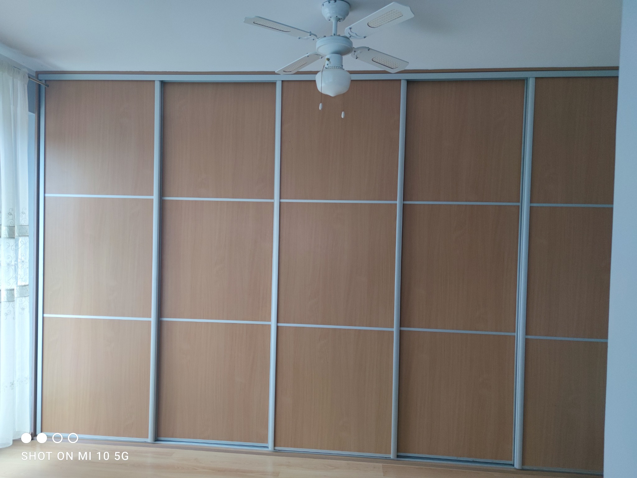 Duża szafa przesuwna z jasnego drewna z szarymi metalowymi ramami, zajmująca całą ścianę, z widocznym wentylatorem sufitowym z lampą pośrodku.