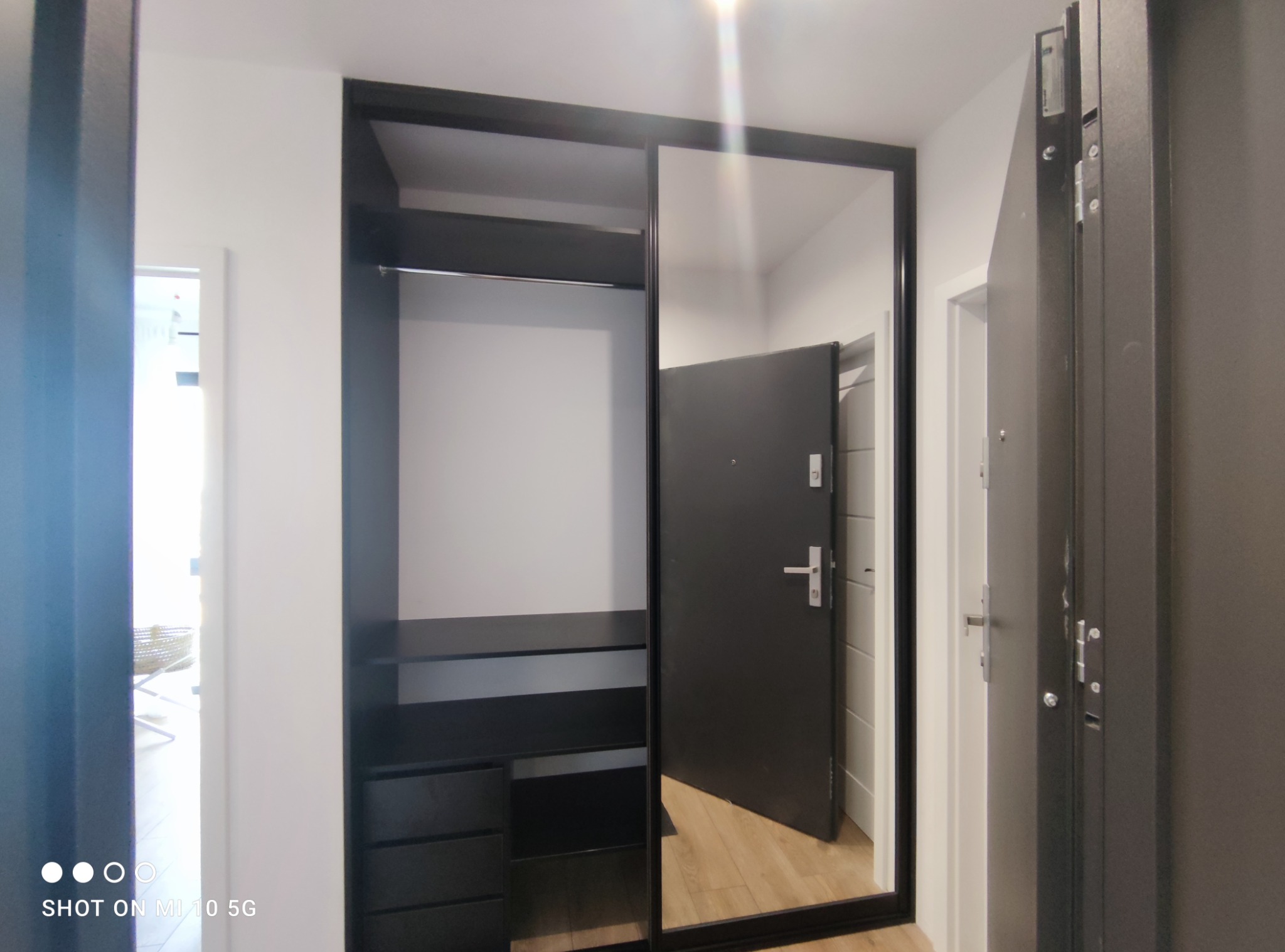 Czarna szafa przesuwna z lustrem w przedpokoju, widoczna półka i szuflady, w lustrze odbija się otwarte drzwi wejściowe.